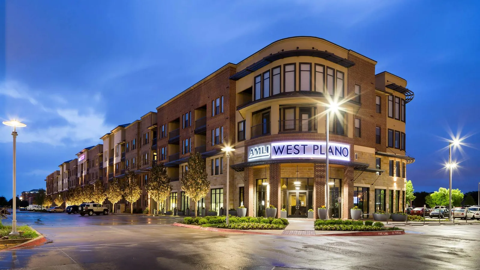 AMLI West Plano in Plano: four-story brick apartment building with ground-floor shops and a lit AMLI West Plano sign on its rounded corner at dusk