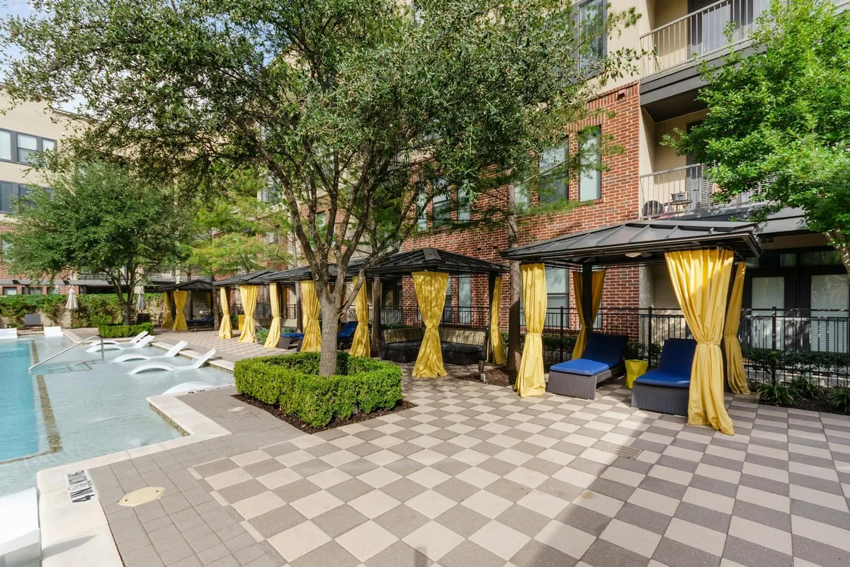 AMLI West Plano in Plano: pool deck with checkered pavers, cabanas with yellow curtains and loungers under mature trees beside a brick building