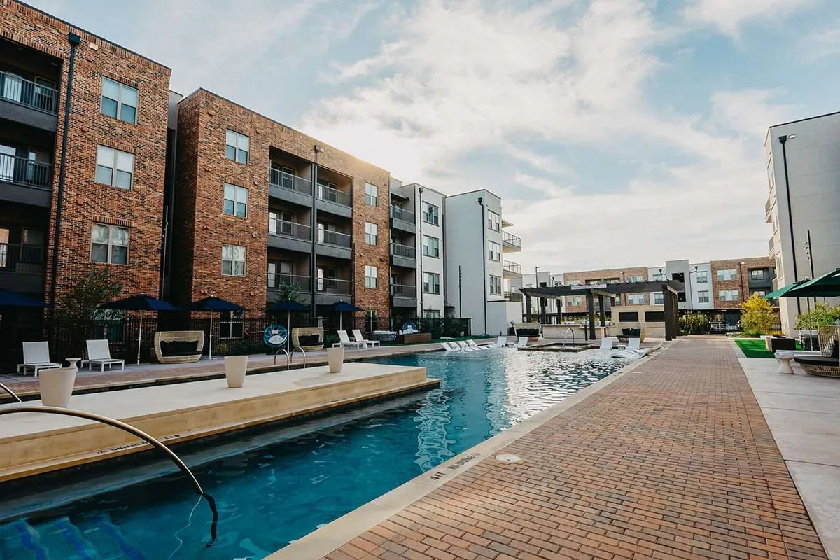 Arden at Midtown GP in Grand Prairie: long pool beside a brick walkway, with brick apartment buildings and green umbrellas along one side