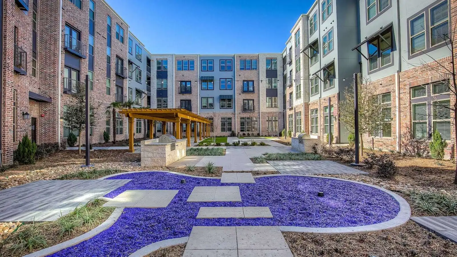 Cole Park in McKinney: landscaped courtyard with a stone path through blue glass mulch and a wood pergola, framed by four-story brick apartment buildings