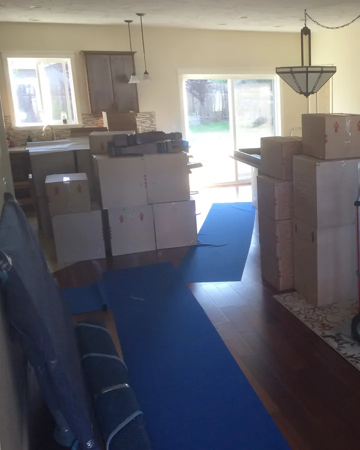Moving boxes stacked between the kitchen and a sliding patio door, with blue floor protection and a rolled-up moving blanket in front