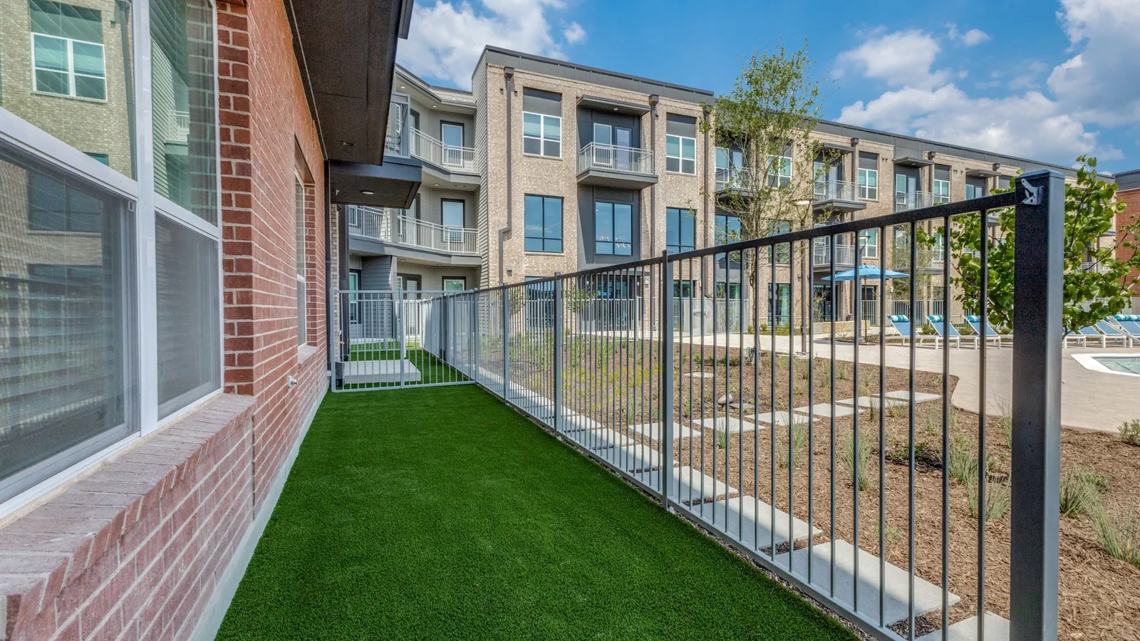 Jefferson North Ranch in McKinney: ground-floor patio with a private artificial-turf yard and a metal fence beside four-story apartment buildings under a blue sky