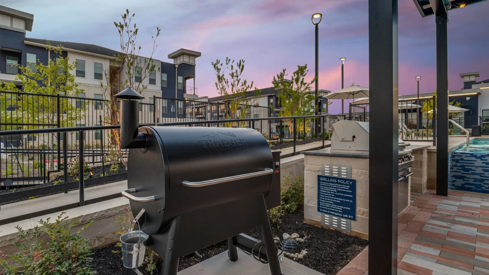 Jefferson Prairie Park in Fort Worth: outdoor grilling area with a black smoker grill and a covered kitchen counter, with apartment buildings behind at dusk