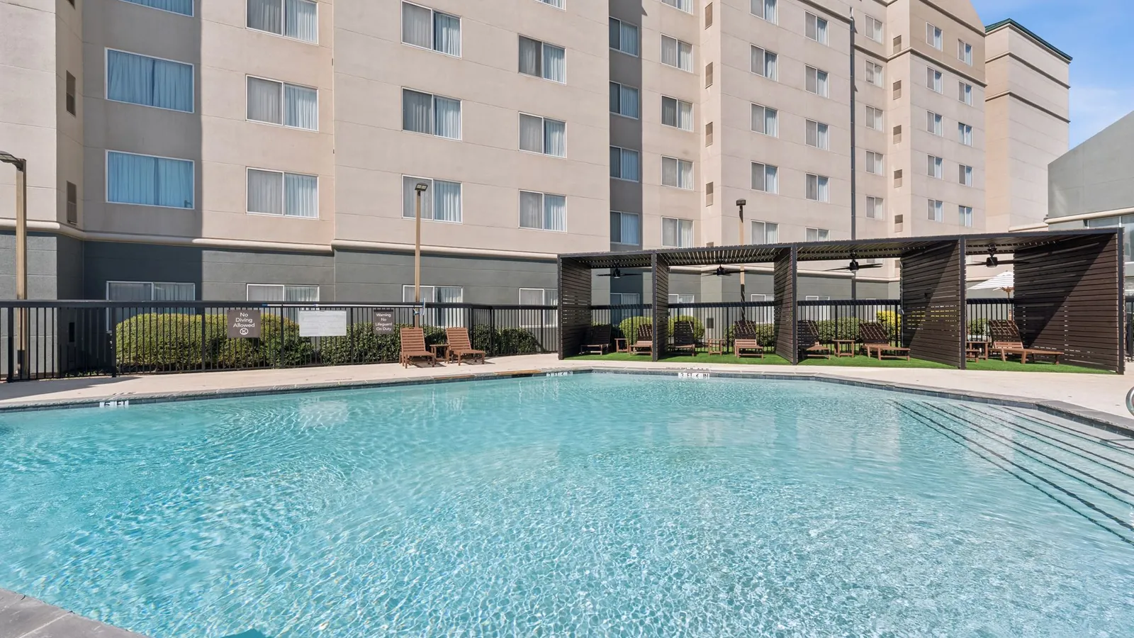 The Ainsley in Dallas: seven-story beige apartment building behind a fenced swimming pool and a shade pergola