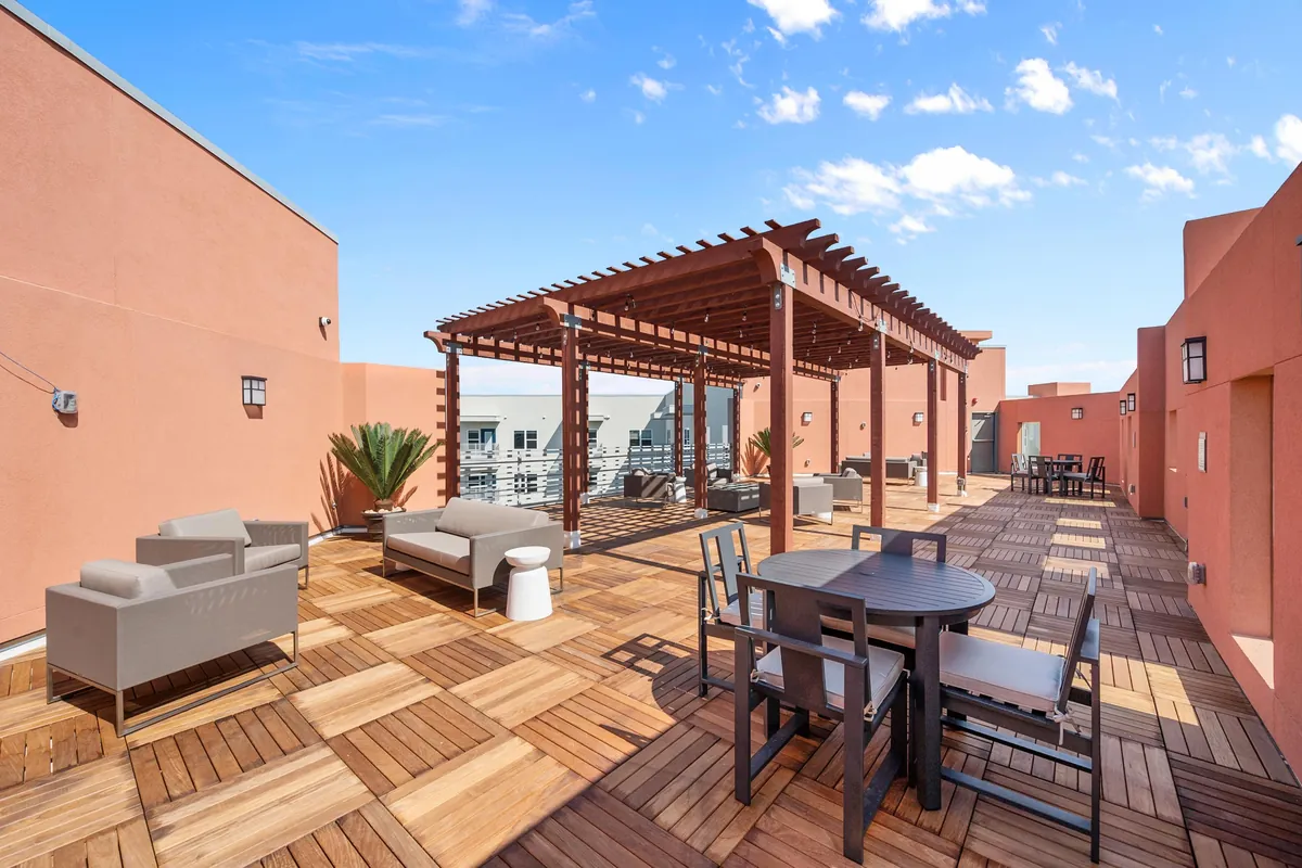 The Gibson in Grand Prairie: rooftop terrace with wood deck tiles, a red pergola, lounge chairs and a dining table between terracotta walls