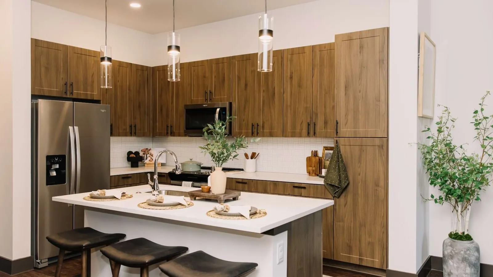 The Harden at Public Market in Fort Worth: kitchen with wood-grain cabinets, a white island with black stools, glass pendant lights and a stainless refrigerator