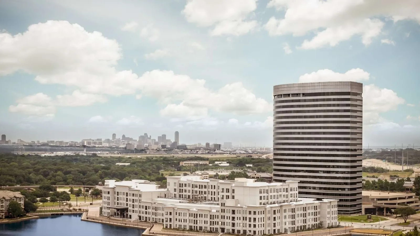 The Mustang in Irving: aerial view of a white mid-rise apartment building beside a lake, with a tall round office tower and a city skyline on the horizon