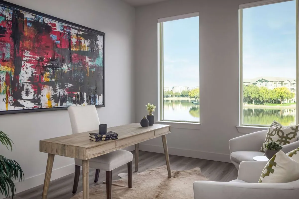 The Mustang in Irving: living area with a wood desk, a white armchair, a large abstract painting and tall windows facing a lake