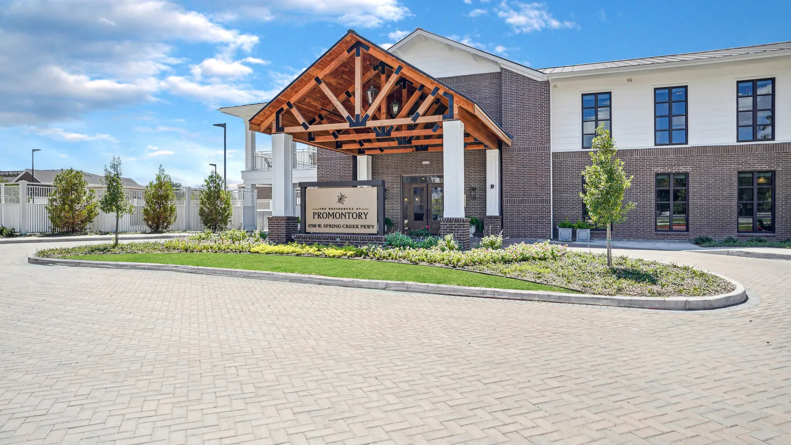 The Residences at Promontory in Plano: two-story brick and white leasing building with a timber-framed entry canopy over a paver drive and a lawn