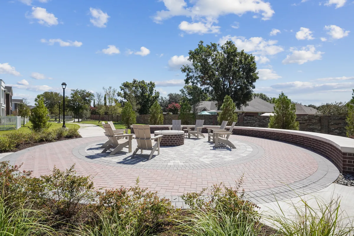 The Residences at Promontory in Plano: circular brick-paved fire pit plaza with Adirondack chairs and low seating walls, under a blue sky with clouds